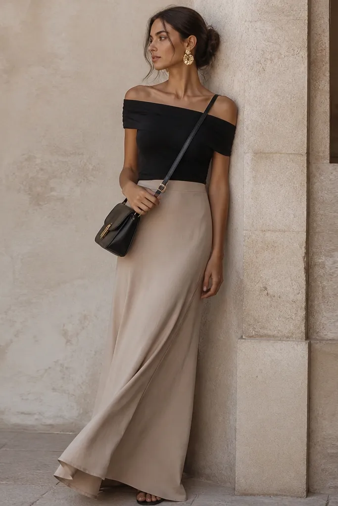 A stone-beige maxi skirt with a flowing hem. A black off-shoulder top sits across the shoulders. Large gold statement earrings and a black crossbody bag are shown.