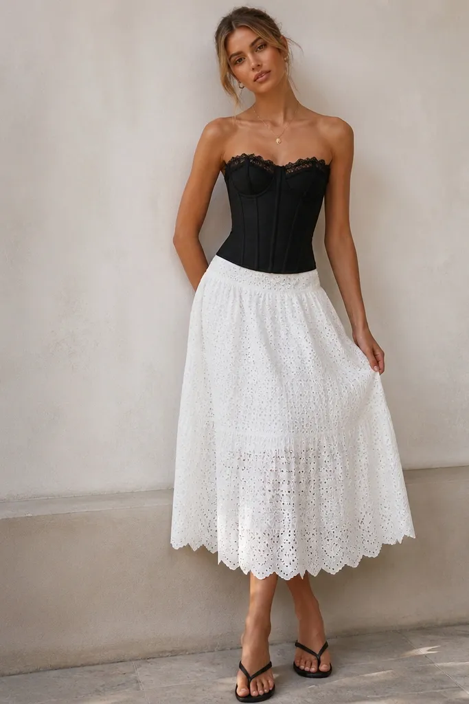 A black corset with lace trim is paired with a white eyelet skirt that has a scalloped hem. The model wears a delicate chain necklace and flat sandals. The image shows a high corset placement and a textured white skirt.