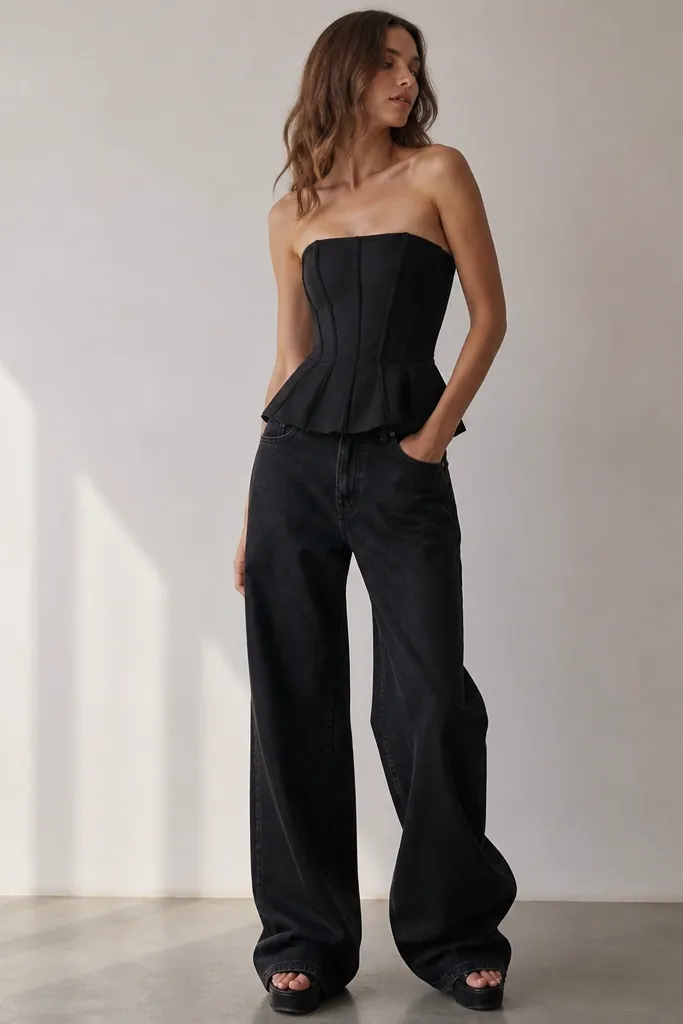 A black corset top with a peplum seam at the waist and structured boning. Black wide-leg jeans drape loosely and pool slightly at the ankle. The outfit includes platform sandals.