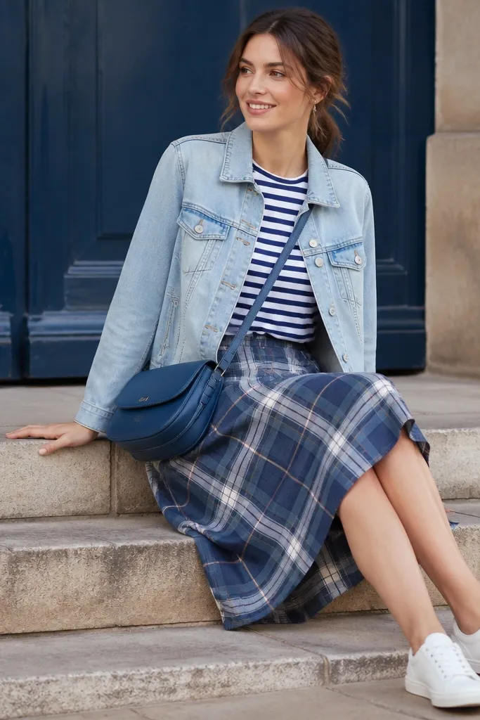 A faded light-wash denim jacket worn open over a navy-and-white striped tee. The outfit pairs with a knee-length blue plaid skirt. White sneakers and a blue crossbody bag.