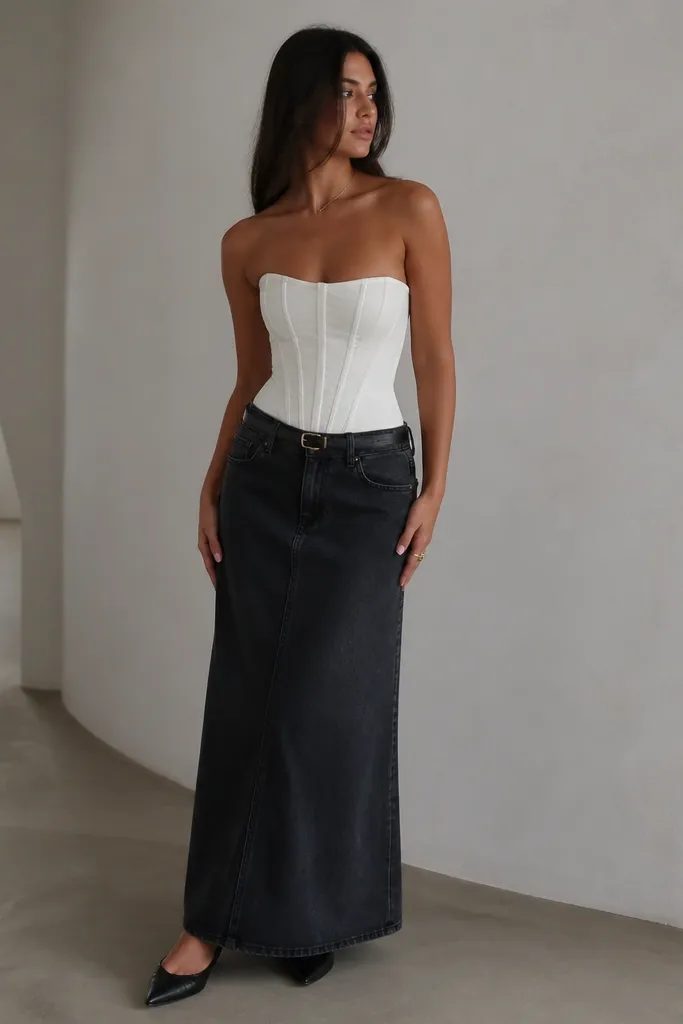 A white corset-style bodice top with boning lines and a fitted silhouette worn over a black denim maxi skirt. The model wears black pointed-toe flats and a slim black belt at the waist.