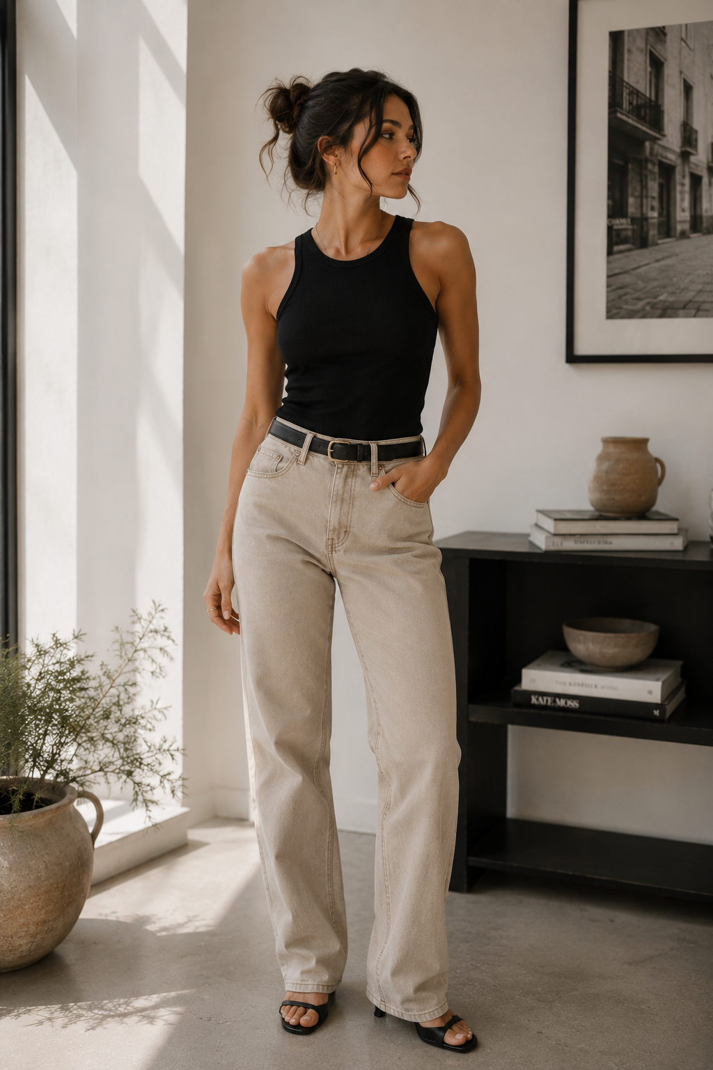 Stone-beige chinos/jeans style pants with a black tank top. A black belt with a simple rectangular buckle is visible at the waist. The model wears dark brown loafers and has a neat watch on the wrist.