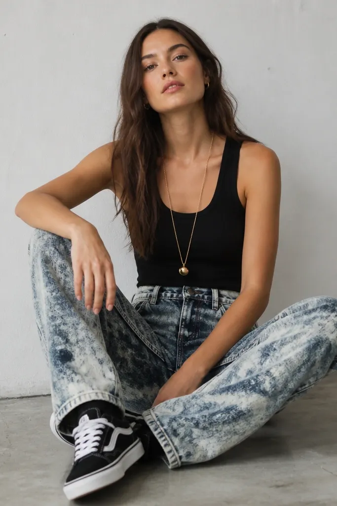 Acid-wash jeans with a high-contrast faded pattern. A black tank top sits cleanly at the waist. A long gold necklace hangs down the center, and the model wears black and white sneakers.