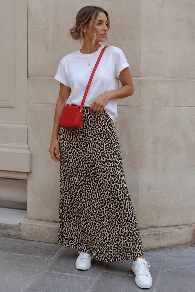 A plain white short-sleeve tee tucked at the front third into a leopard print maxi skirt. A bright red crossbody bag stands out. The footwear is white sneakers.